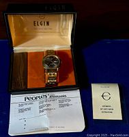Elgin wristwatch displayed in its original black and orange box with the watch centered and the open lid showing 'Elgin Swiss Made Since 1864'. Below the box is a folded document from Peoples Jewellers.
