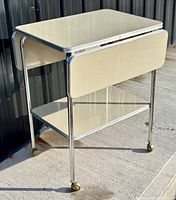 Photo of the vintage 1950s formica drop-leaf bar cart showing one drop leaf down and the other up, on gold casters.