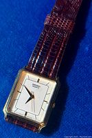 Close-up photo of the gold tone rectangular watch case with silver dial and brown textured leather strap, showing the dial and watch hands.