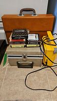 Stacked assorted 8-track tapes on top of silver electronic player case with nearby yellow device, tan carrying case behind
