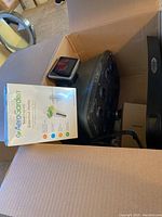 Cardboard box with a black AeroGarden base unit inside along with a packet of AeroGarden Gourmet Herbs seeds.
