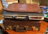 Photo showing stacked items: a brown leather document case on top, a black briefcase in the middle with metal edges, and a dark brown leather textured briefcase at the bottom.