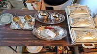 Overview photo showing a variety of silver-tone serving pieces on a wood table including cream and sugar sets, serving platters, and small stackable plates