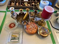 Various brass and ceramic candle holders, red and white pillar candles, clear glass ashtrays with decorative elements, ceramic bowl with marine motifs, and a brass incense burner with enamel decoration.