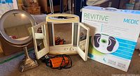 Shows the Revitive Circulation Booster in box, the tri-fold illuminated make-up mirror, and the swiveling pedestal mirror together on floor.