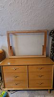 Full view of a light wood dresser with a separate rectangular mirror frame placed on top, shown against a textured wall with some wear visible on surfaces.