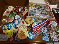 Wide shot showing a large assortment of pins and buttons scattered on a table, showcasing various holiday, event, and souvenir designs including Canadian maple leaf symbols and novelty logos.
