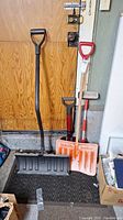 Group photo of three snow shovels and one vehicle broom against a wooden door.