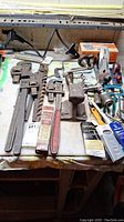 Overview of assorted tools laid on a table, showing multiple pipe wrenches, an auger drill bit, paint brushes, snips, and hand planes.