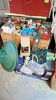 Photo showing multiple boxes with vintage Christmas decorations including boxed glass ornaments, string lights, and a green metal tree stand on the floor.