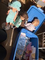 Three vintage dolls standing next to a blue storage bin containing doll clothing.