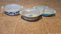 Three vintage Pyrex casserole baking dishes with varying patterns (black floral/fruit, brown floral, blue snowflake) with glass lids placed on tile floor.