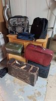 Photo showing several vintage suitcases and bags of various materials and colors: black, silver tote, red leather, green metal box, and worn brown suitcase.