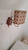 Side angle of nautical ship's wheel wall lamp mounted on white wall, showing fabric shade and pull chain. Power cord visible running down wall.