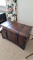 Wooden dark brown steamer trunk with metal clasp and decorative leather-like straps with metal studs, shown from angled top side view.