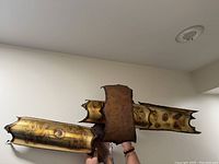 Photo showing the sculpture held by hands against a plain wall, displaying the brass and copper material with a rich patina and rough edges.