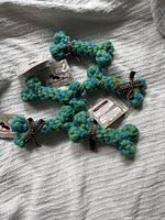 Top view of 5 blue and green natural cotton braided dog bone toys with brown ribbons on a fabric surface, tagged brand new.