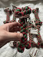 Close-up of a hand holding one of the red and green braided cotton dog bone toys with attached tag and brown ribbon.