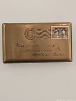 Front view of brass cigarette case resembling a mailed envelope with address and Canadian stamp details.