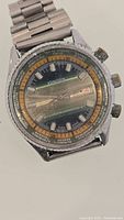 Front view of the vintage Kronotron World Time mechanical watch showing the gradient green-orange dial, date window at 3 o'clock, three crowns on the case edge, and attached steel bracelet.