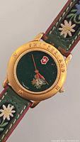 Close-up view of watch face showing gold-tone bezel engraved with 'SWITZERLAND', green dial with floral design and Swiss emblem, gold and red hands, and the original embroidered green leather strap with floral stitching.