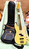 SX Vintage Series electric bass guitar with pale yellow body and white pickguard leaning against blue padded soft case with multiple zippered compartments and brown trim, also shows a black Ernie Ball strap attached to the guitar.