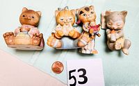 Four glazed cat figurines shown from front, including one with yarn, one in pajamas holding doll, one holding bottle, and one sleeping, arranged on light fabric with penny for scale.