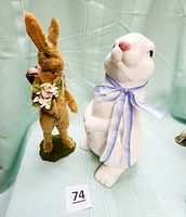 View of both the tall brown fuzzy rabbit holding a bouquet and the ceramic white bunny with a blue bow.
