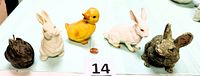 Front view of five figurines including two white rabbits, a yellow duckling, a gray and a dark brown bunny on a light blue cloth with size scale and a penny for size reference.