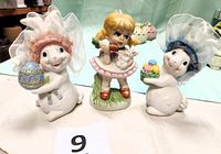 Three porcelain figurines: two rabbits with organdy hats (one pink, one blue) and a girl figurine holding a bunny and carrots, displayed on a white surface with a green background.