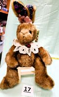 Front view of the brown plush bunny sitting on the painted wooden chair. Shows the bunny's fur, lace collar, floral inner ears, and pink bow.