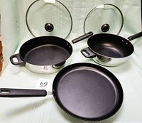 Three Cook's Essential pans arranged with lids behind two pans. The 10-inch and 11-inch pans have glass lids, black handles, and an additional safety carry handle opposite the main handle. The 12-inch skillet pan has a single black handle and no lid.