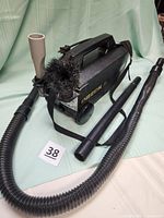 Oreck XL black compact canister vacuum with carrying strap and flexible hose coiled around, viewed from the side showing the attachments on-board.
