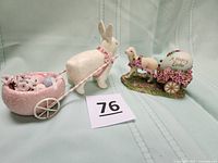 Photo showing two Easter-themed figurines: a white rabbit pulling a pink, egg-shaped flower cart with pastel eggs and a white sheep pulling a flower cart with an egg labeled 'A Happy Easter'. Both figurines have glitter detailing.