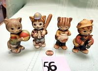 Front view of all four cat figurines showing different sports-related items each is holding, with a penny for scale and lot number 56 visible.