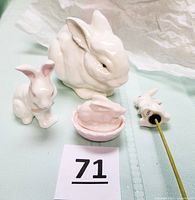 Set of four porcelain rabbit figurines including a large white rabbit, a small white rabbit with pink ears and bow, a pink bunny on basket nest ring holder, and a bunny on a stick for decoration.