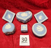 All six miniature Wedgwood Jasperware trinket boxes and dishes arranged on red fabric, showing tops and variety of shapes distinct to Wedgwood style with characteristic pale blue and white colors.