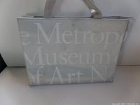 Full front view of the beige tote bag with white Metropolitan Museum of Art printed text and leather handles.