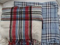 Two wool-blend plaid scarves or wraps, one cream-white with red and black plaid patterns and the other blue plaid pattern, both folded with fringed ends visible.