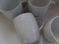 Four white ceramic mugs each embossed with a large snowflake motif on the front side, arranged with one tilted forward showing the design clearly.