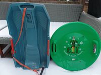 Pair of sleds: blue brake sled showing the interior and red pull strap, green round Buddy the Elf saucer sled showing top design and handles.