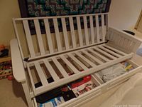 White wooden double futon frame with slatted armrests and backrest, open to show mattress support slats and storage drawers underneath filled with miscellaneous household items.