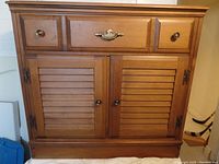 Front view of the solid maple cabinet showing central drawer with decorative brass handle, louvered double doors below, two smaller drawers, and round brass knobs.