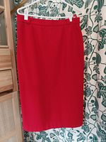 Full view of the red midi pencil skirt hanging on a white hanger against a background with green floral pattern, showing the straight tailored shape and length.