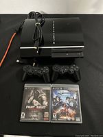 Sony PlayStation 3 console with power cord, two black controllers, and two video game cases on black surface.