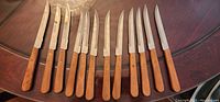 Ten serrated steak knives with stainless steel blades and wooden handles laid out in a row on a dark wooden table.