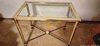 Gold metal rectangle shaped end table with glass top, showing overall front view including the metal crossbar brace and round legs.