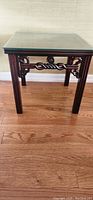 Single carved rosewood end table with a glass top, shown on a hardwood floor against a wall.