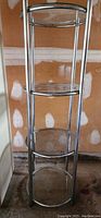 Full view of a round 4-tier glass display stand with a tubular metal frame, placed indoors near a wall.