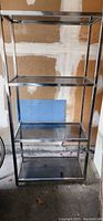 Glass and metal display stand with four glass shelves and metal frame, shown standing against a wall with a pegboard in the background.
